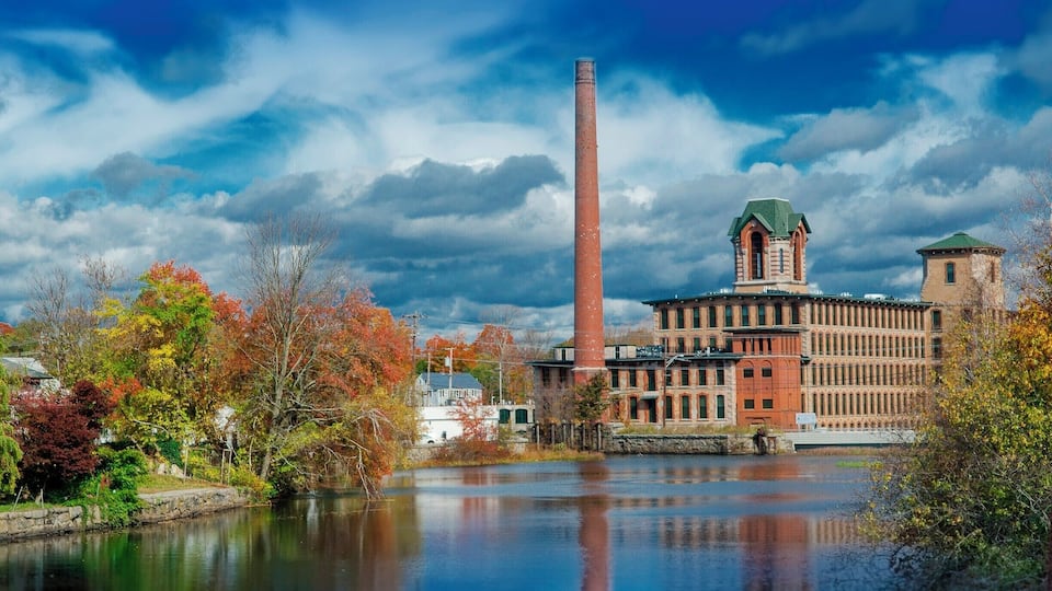 A historic building with a tall chimney, a river, and autumn foliage.