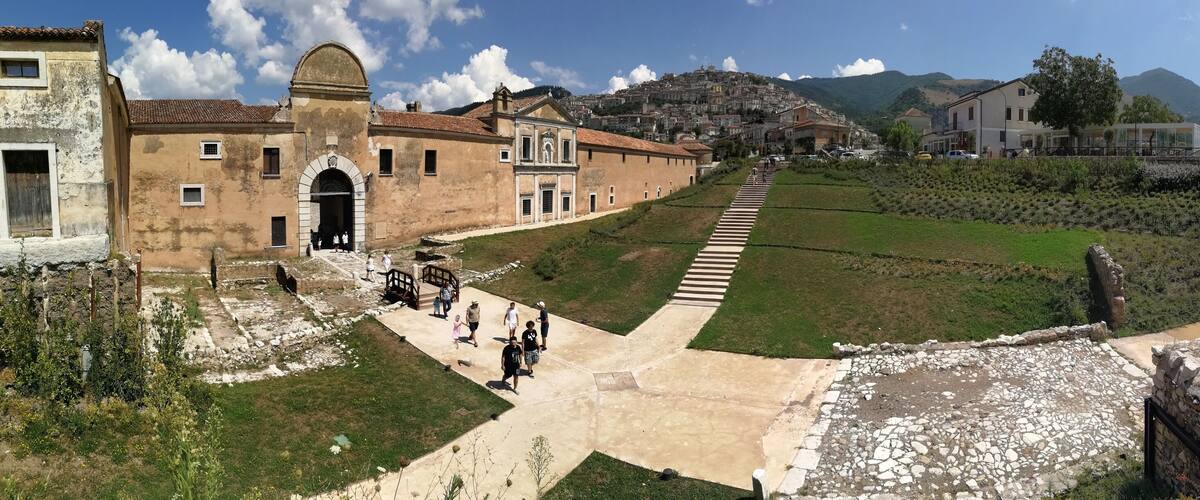 A historical site with ancient buildings, a paved pathway, and a hillside with a small settlement in the background.