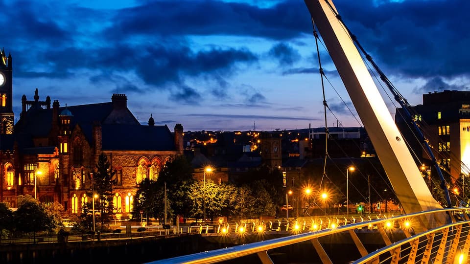 A cityscape at dusk with a prominent clock tower, a modern bridge, and illuminated buildings.