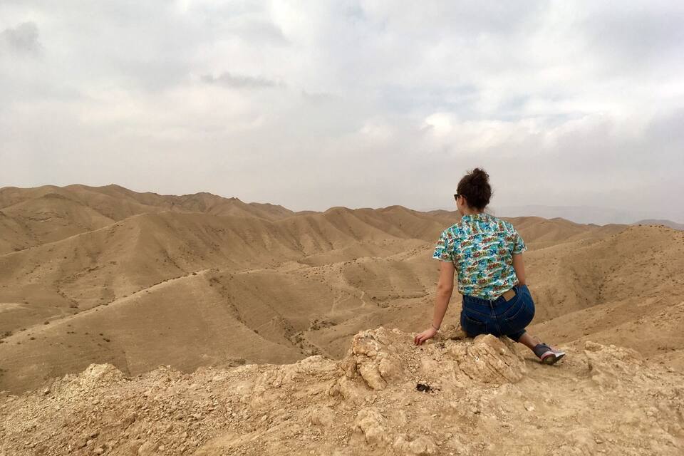 A person sitting on a rocky outcrop overlooking a vast desert landscape with undulating sand dunes.