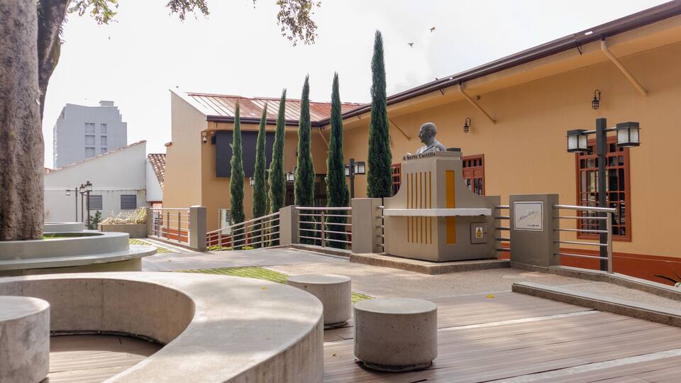 A courtyard with a statue, tall cypress trees, and a building with a yellow facade.
