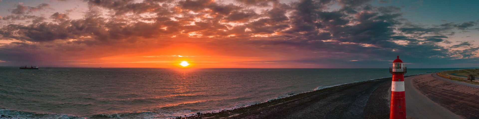 A sunset over the ocean with a red and white lighthouse on a coastal road.
