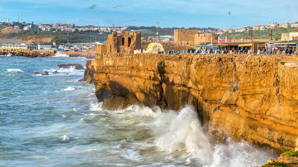 A coastal cliff with waves crashing against it, a historic fort, and a town in the background.
