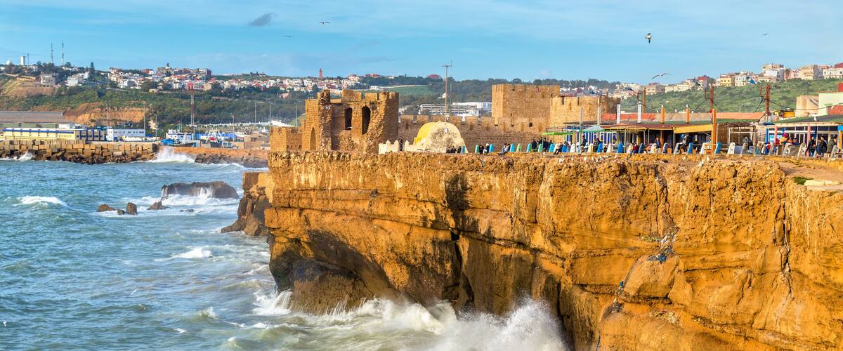 A coastal cliff with waves crashing against it, a historic fort, and a town in the background.