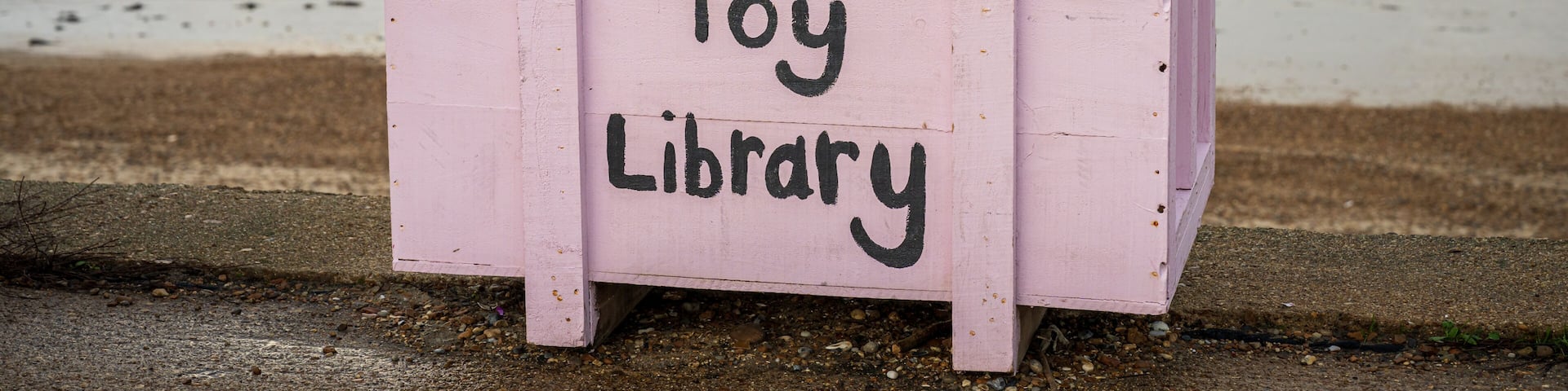 A pink wooden box labeled 'Toy Library' with toys inside, a metal railing, and a lighthouse in the background.