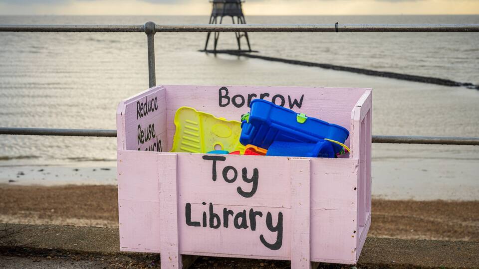 A pink wooden box labeled 'Toy Library' with toys inside, a metal railing, and a lighthouse in the background.
