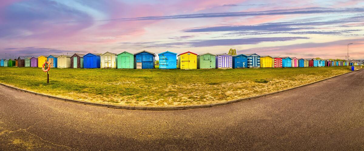 A row of colorful beach huts along a grassy roadside with a sunset sky.