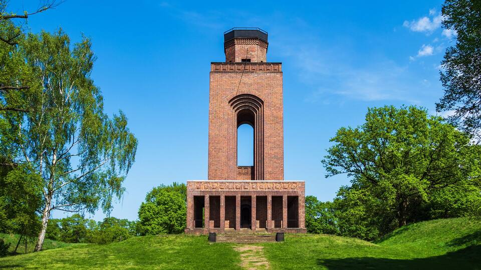 A tall brick tower with a central archway, surrounded by lush green trees and grass.