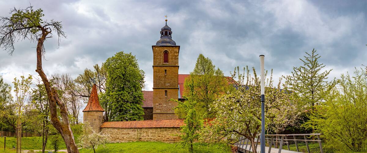 A church with a bell tower, surrounded by greenery and a paved walkway.