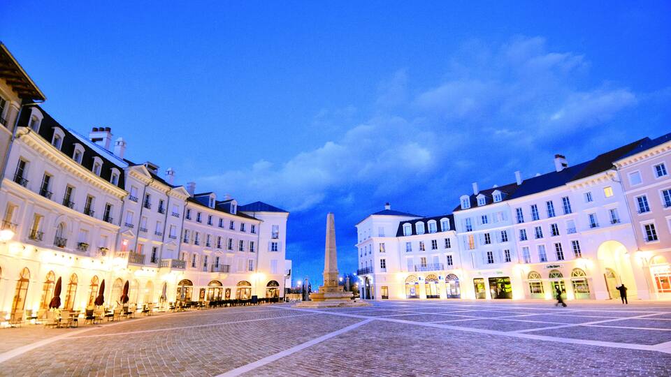A cobblestone square with historic buildings, a central monument, and a clear evening sky.