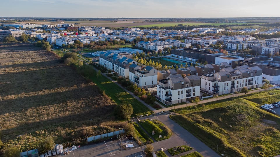 An aerial view of a residential area with modern houses, green spaces, and a distant view of fields and a body of water.