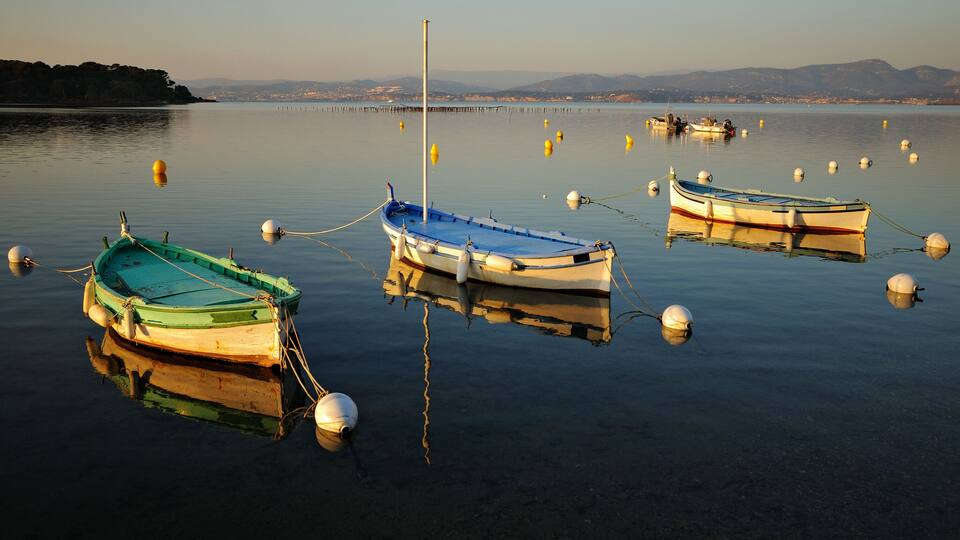 Three boats tied to buoys in calm water with a pier and mountains in the background.