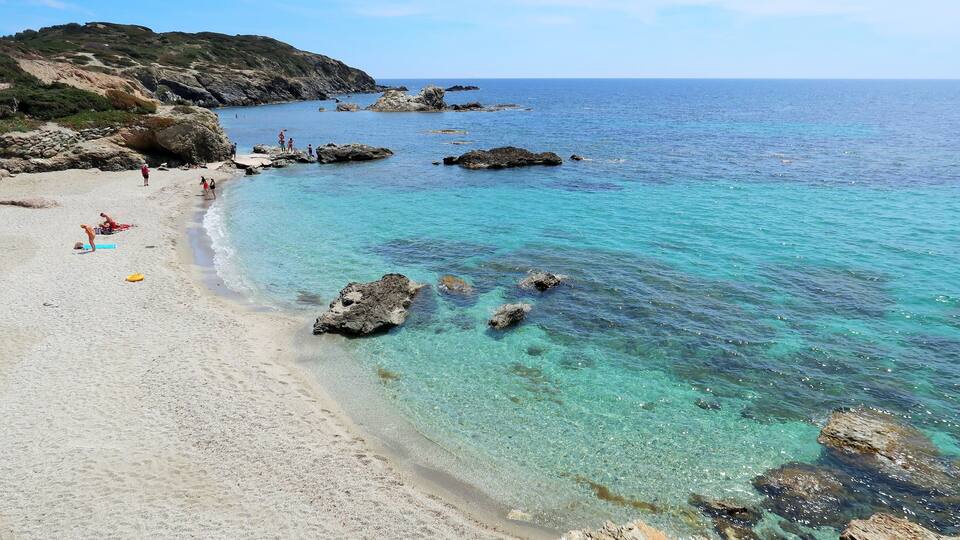 A beach with clear turquoise water, rocky outcrops, and people sunbathing.