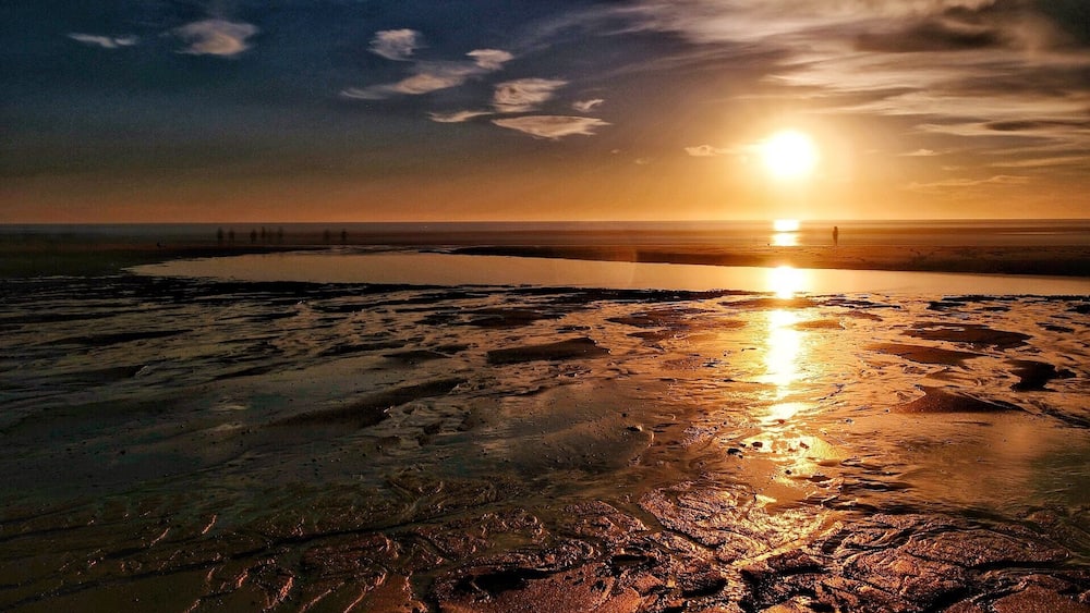 Un coucher de soleil sur une plage humide et scintillante, avec une côte lointaine et une personne debout près du bord de l’eau.