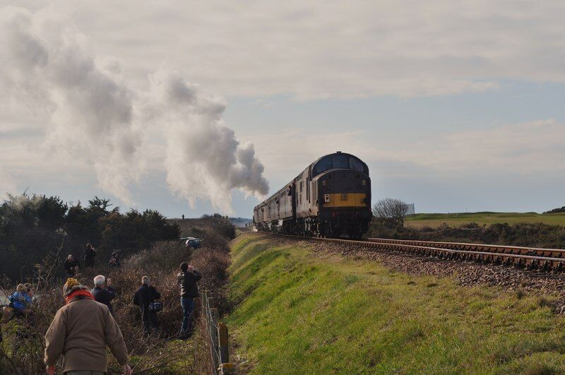 A steam locomotive emitting smoke, passing by a group of people on a grassy embankment.