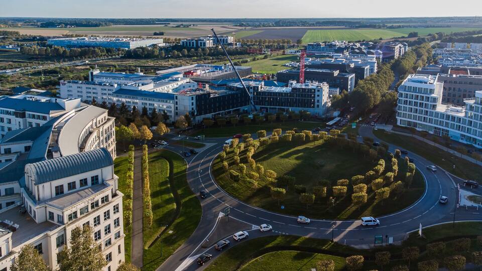 An aerial view of a modern urban area with a central roundabout, surrounded by buildings, roads, and green spaces.