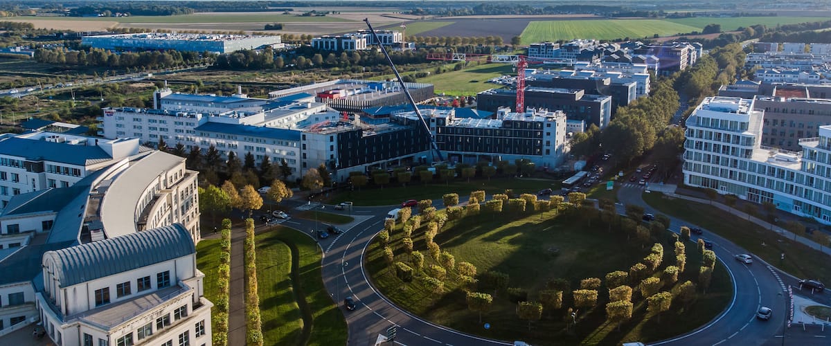 An aerial view of a modern urban area with a central roundabout, surrounded by buildings, roads, and green spaces.