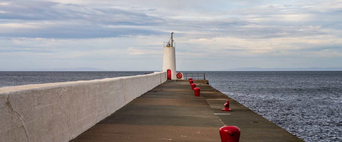 A pier with a lighthouse, red bollards, and the sea in the background.