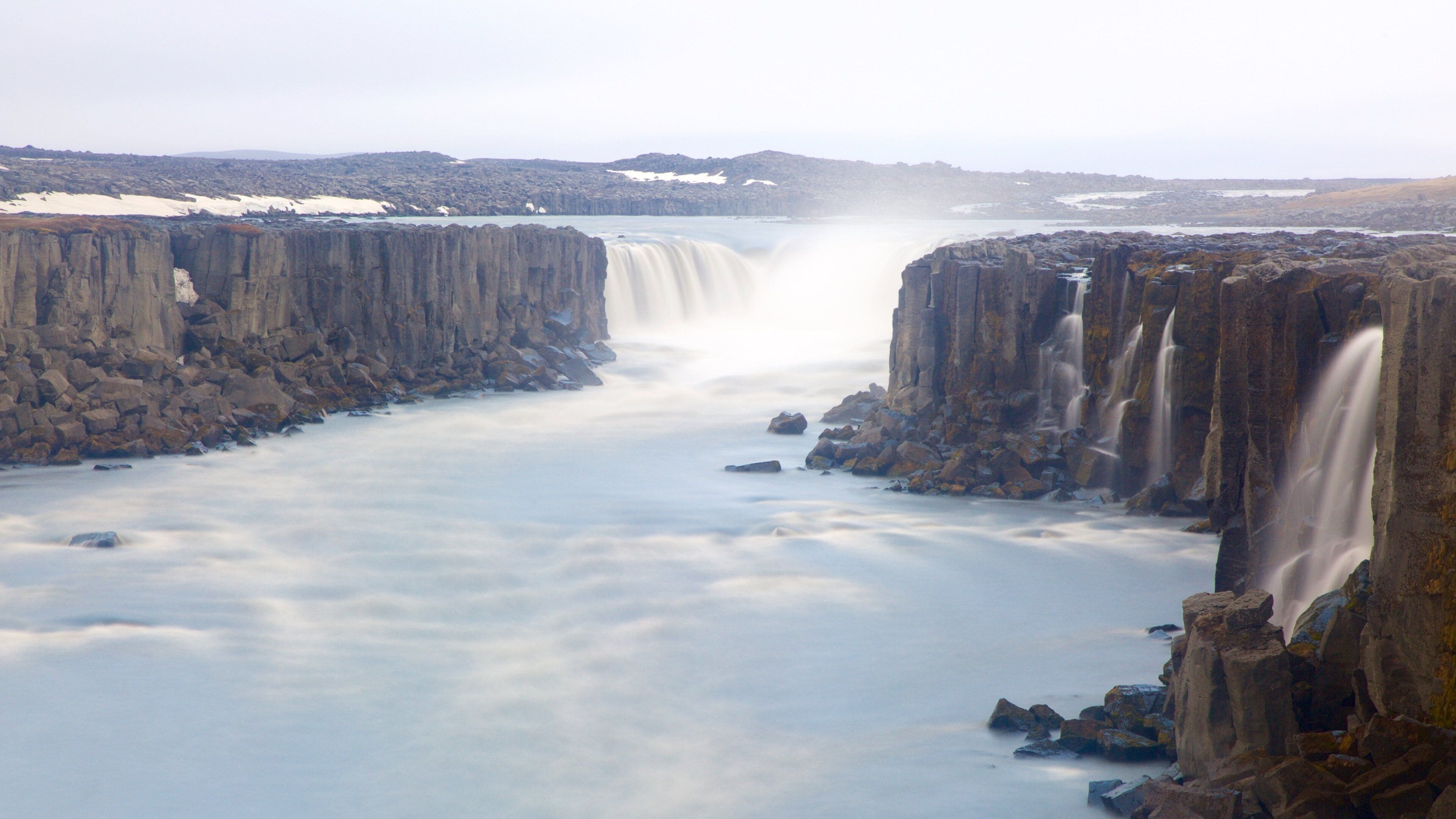 A waterfall cascading down a rocky gorge with snow patches on the surrounding cliffs.