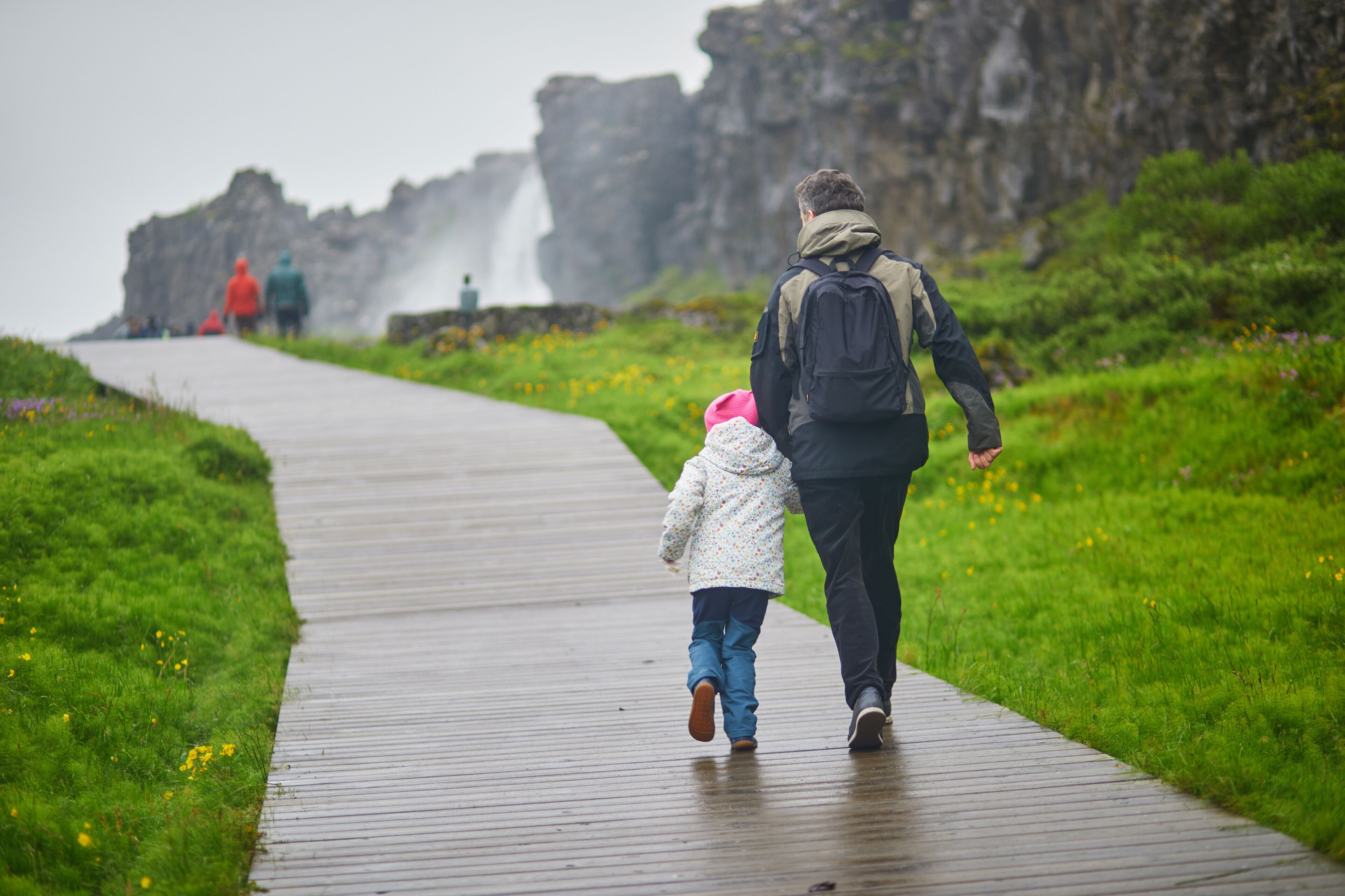 A wet wooden boardwalk with a child and an adult walking towards a waterfall.