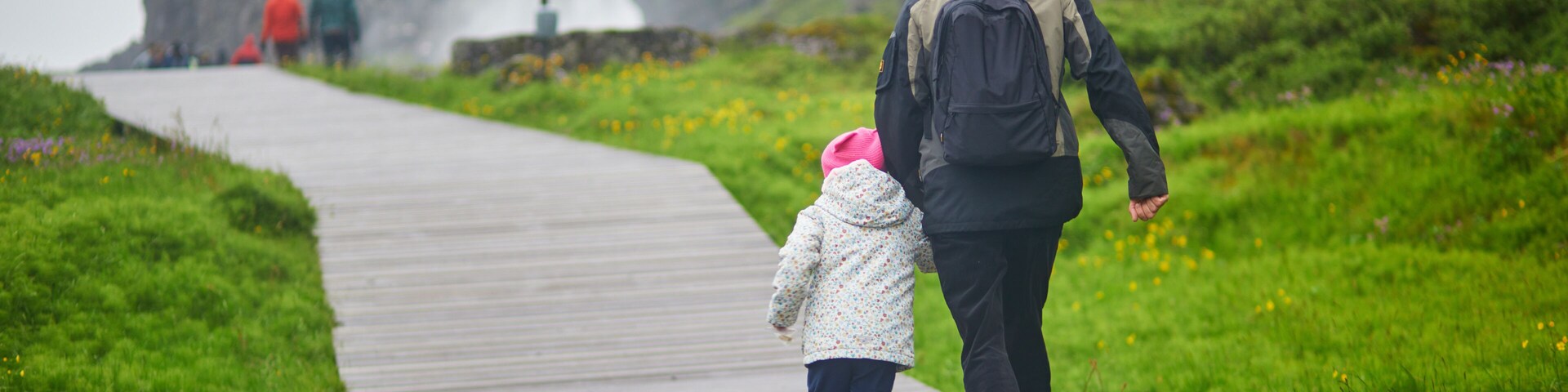 A wet wooden boardwalk with a child and an adult walking towards a waterfall.