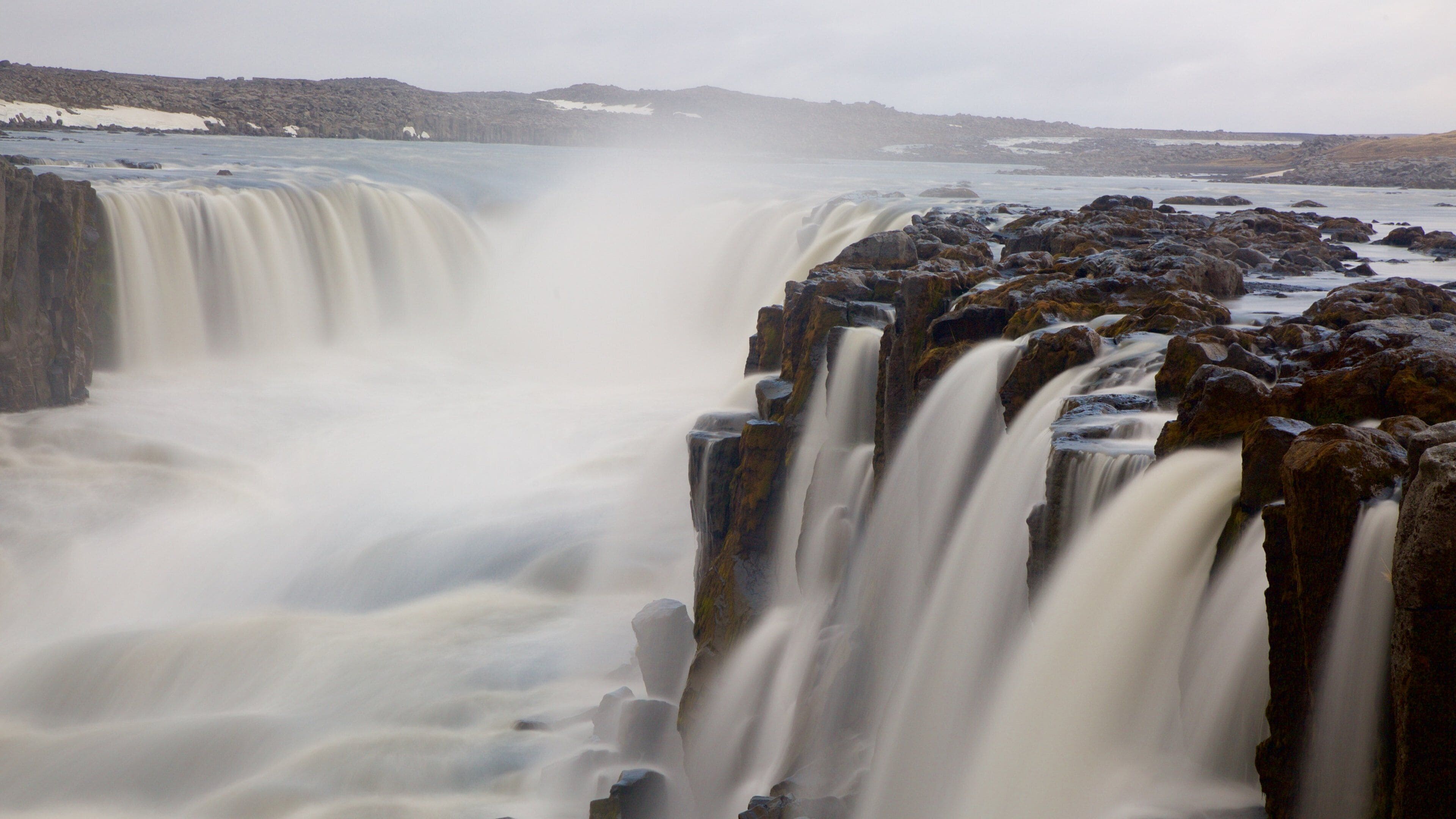 A waterfall cascading over rocky terrain with a distant view of snow-covered hills.