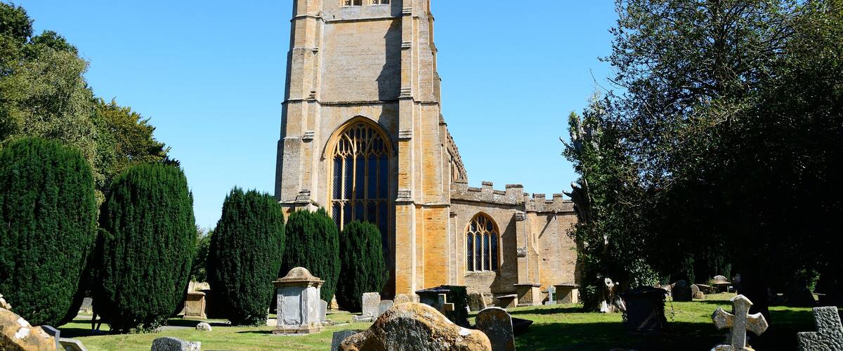 A tall stone church with a clock tower, surrounded by trees and gravestones.