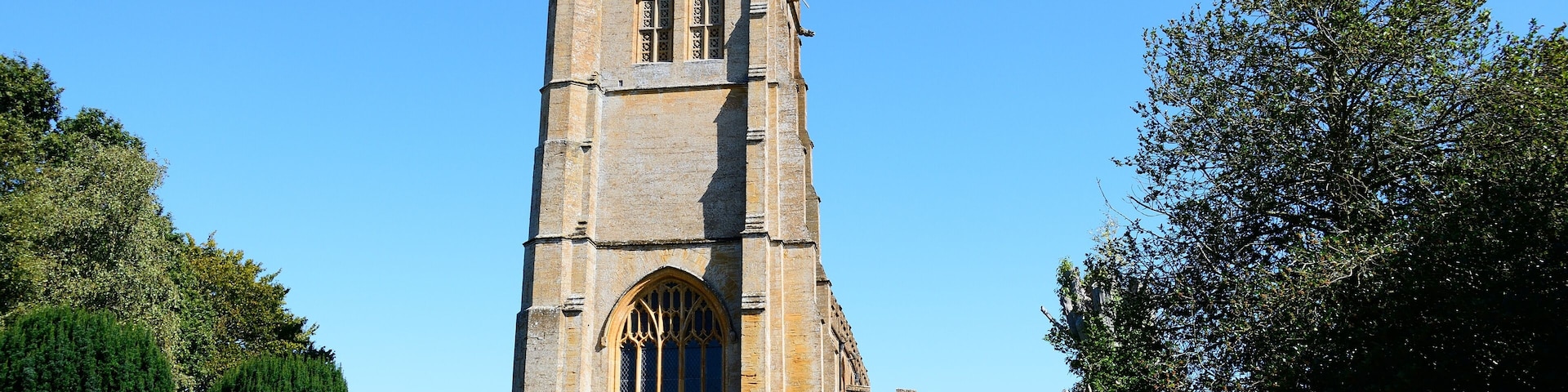 A tall stone church with a clock tower, surrounded by trees and gravestones.