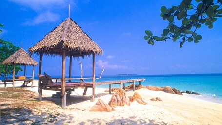 Ein Strand mit strohgedeckten Hütten, einem Holzsteg und klarem, blauem Wasser.