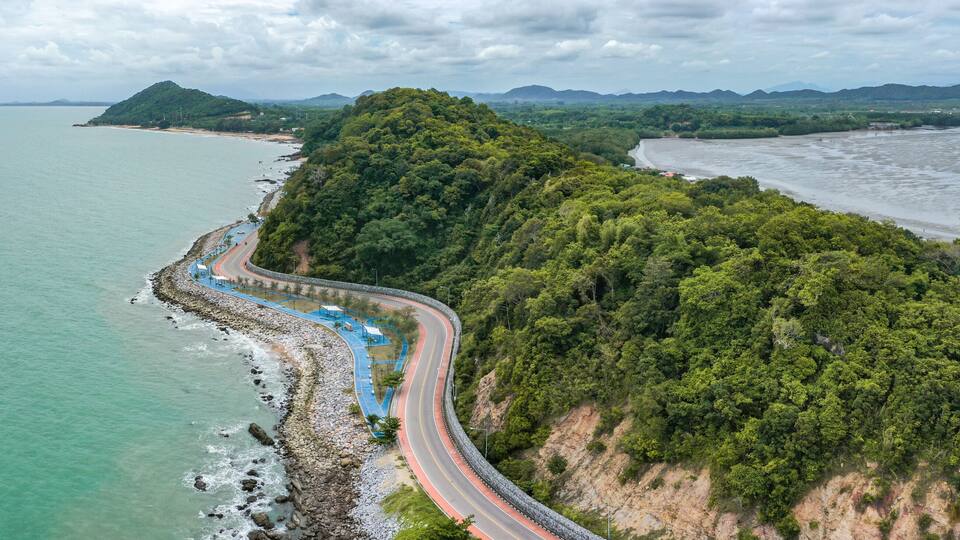 A coastal road winding along a cliffside, overlooking a calm sea and a forested mountain.