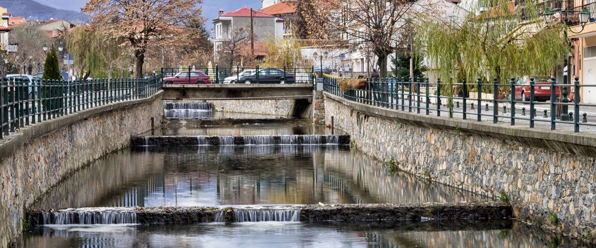 A stone bridge over a calm river with multiple small waterfalls, surrounded by trees and buildings.