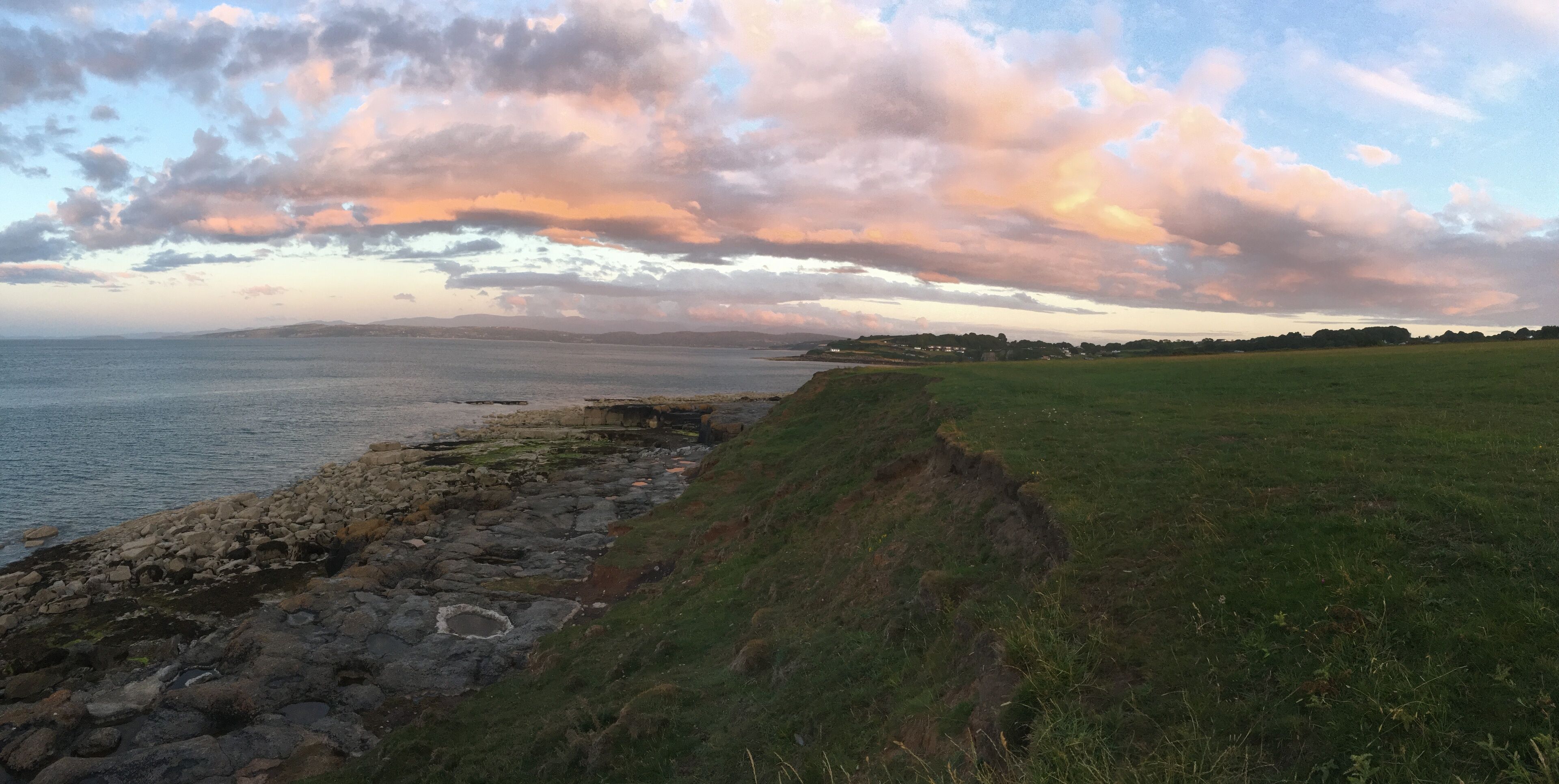 A coastal landscape with a rocky shoreline, grassy fields, and a distant view of hills under a colorful sky.