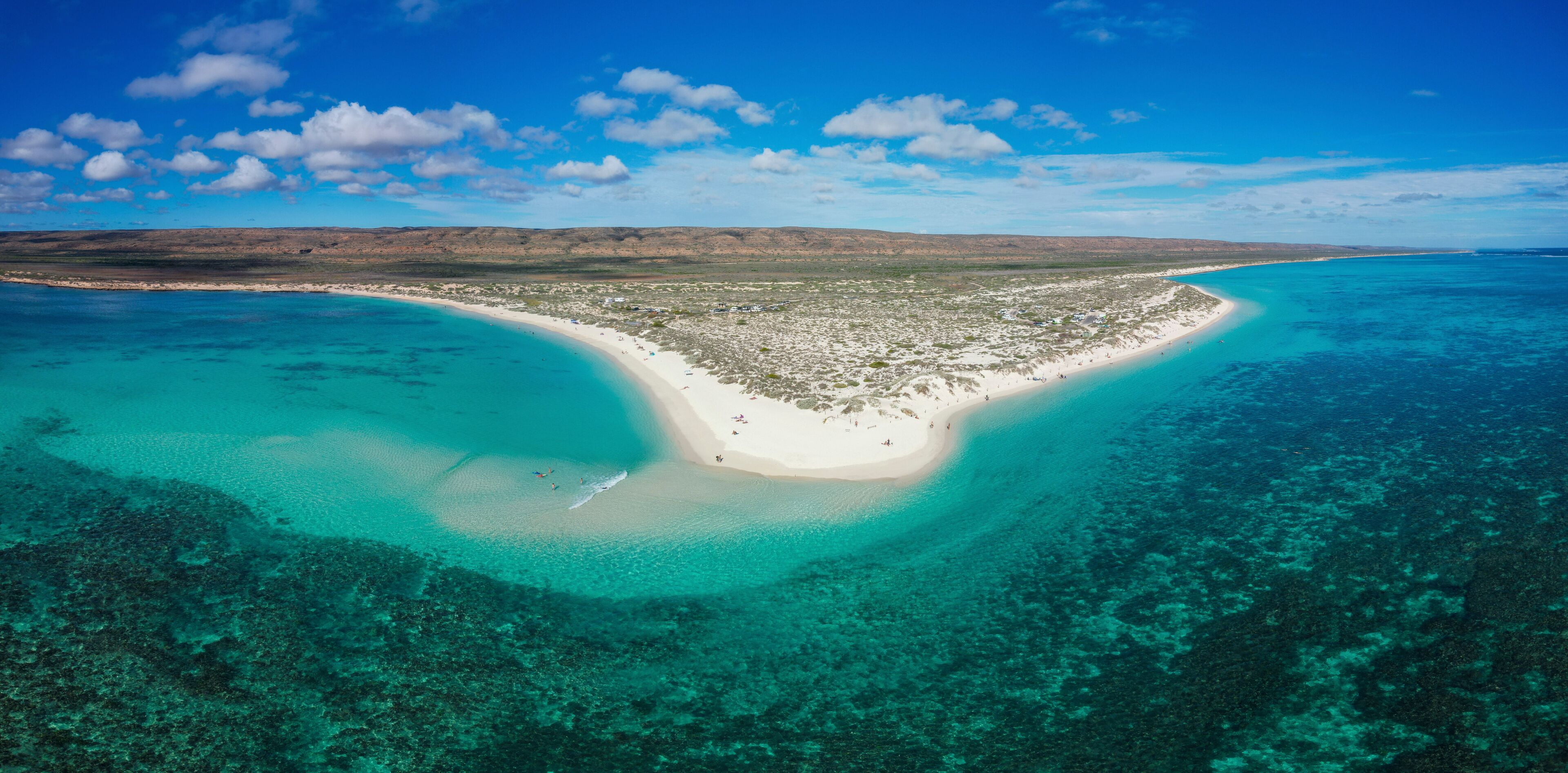 A coastal landscape with a white sandy beach, clear turquoise waters, and a distant mountain range.