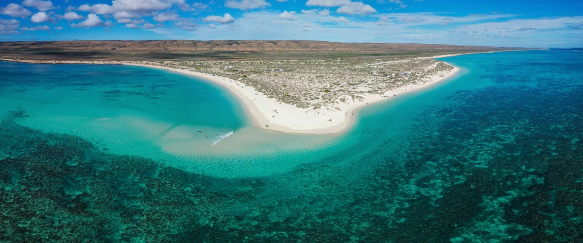 A coastal landscape with a white sandy beach, clear turquoise waters, and a distant mountain range.