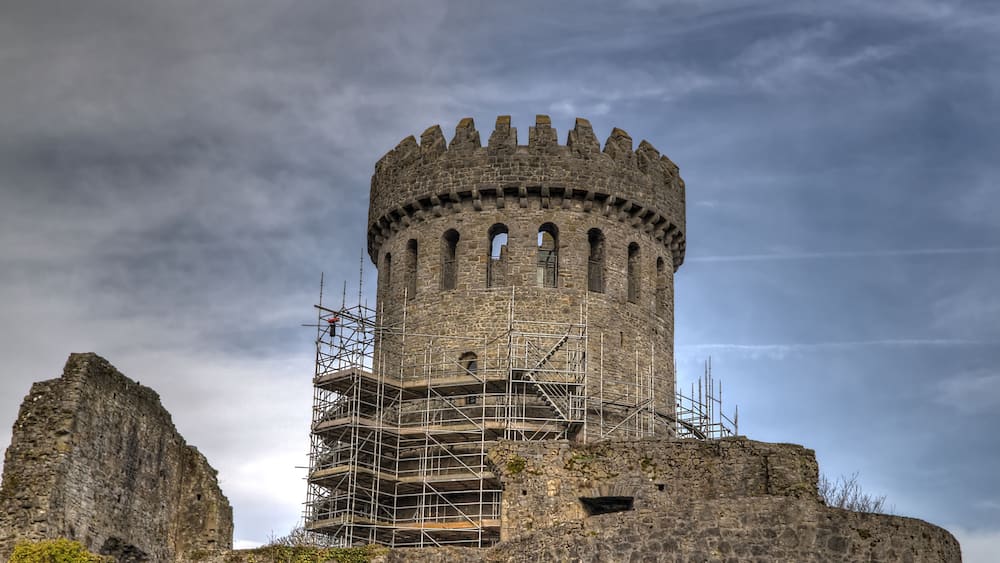 A historic stone tower with scaffolding, surrounded by rugged terrain and a cloudy sky.