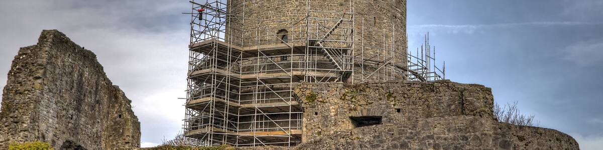 A historic stone tower with scaffolding, surrounded by rugged terrain and a cloudy sky.
