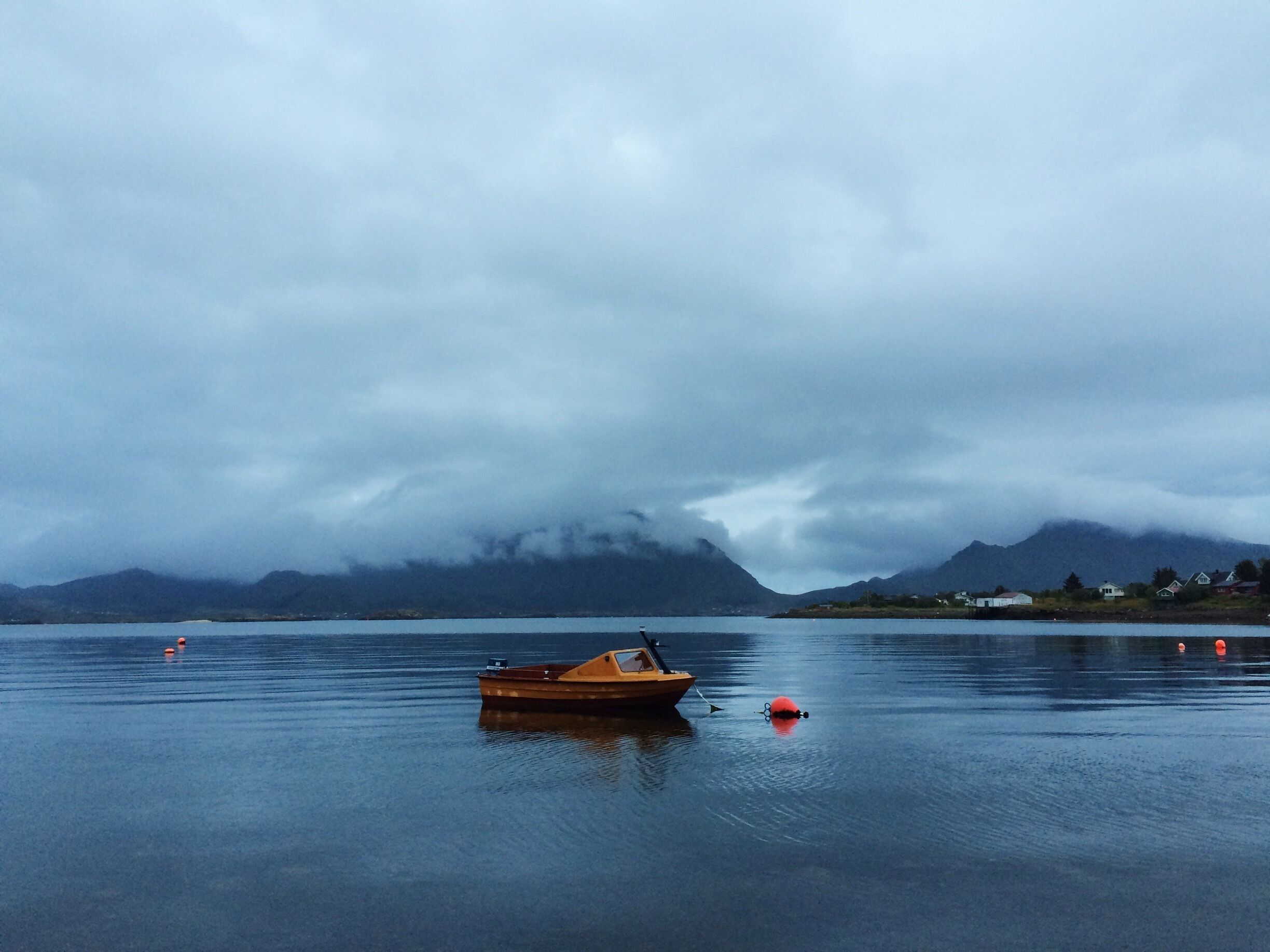 A calm lake with a wooden boat, a red buoy, and a mountain range in the background.