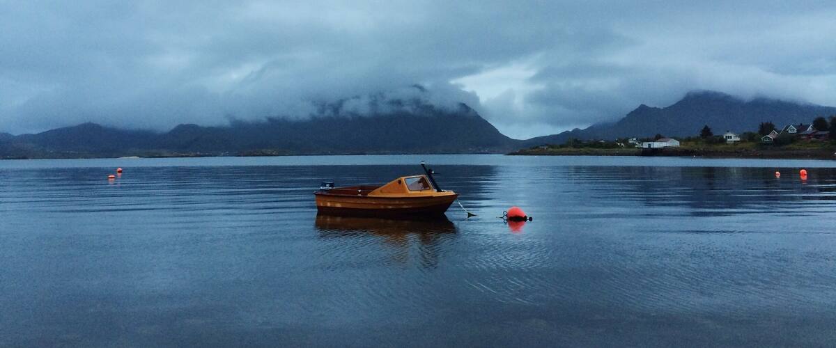 A calm lake with a wooden boat, a red buoy, and a mountain range in the background.