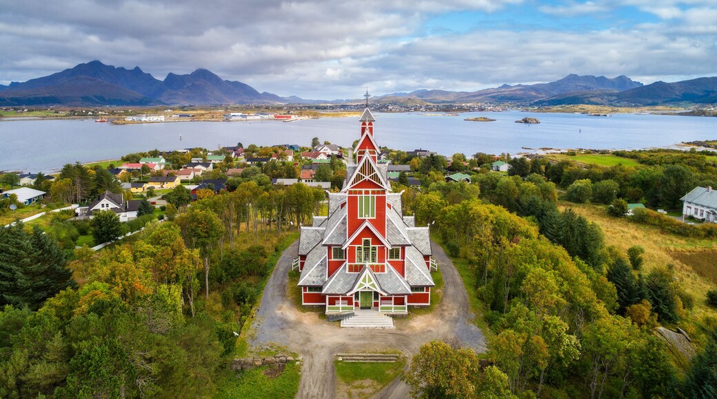 Una iglesia roja con un techo empinado y una cruz en la parte superior, rodeada de exuberante vegetación y un lago con montañas al fondo.