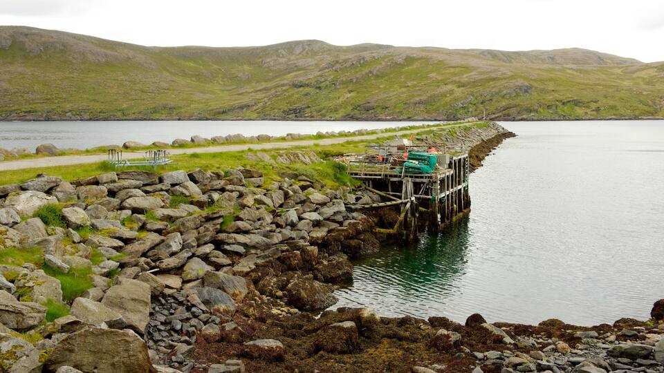 A rocky shoreline with a wooden pier extending into a calm lake, surrounded by green hills.
