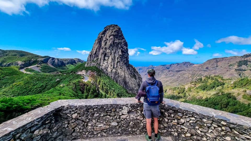 A person standing on a stone wall overlooking a mountainous landscape with a prominent rock formation.