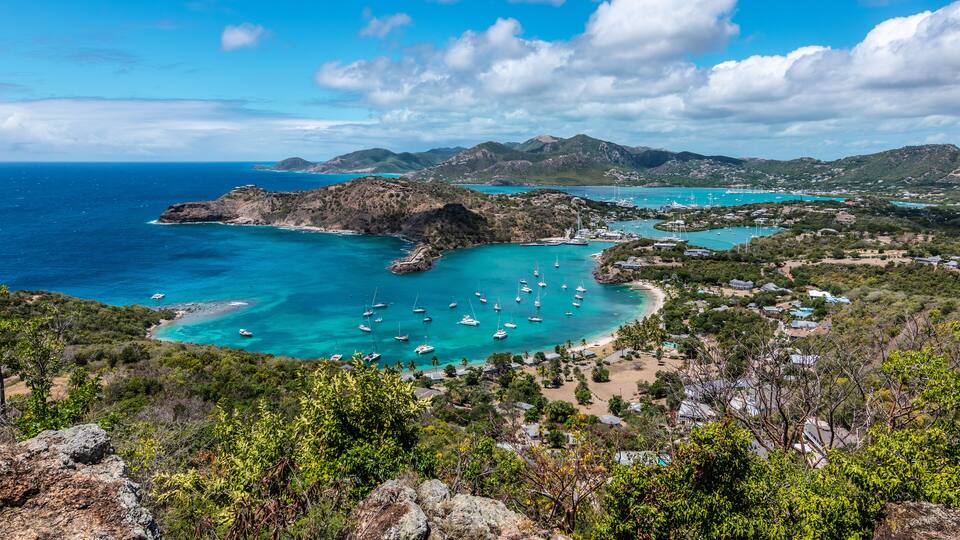 A coastal landscape with clear turquoise waters, a marina with sailboats, and a town nestled among hills.