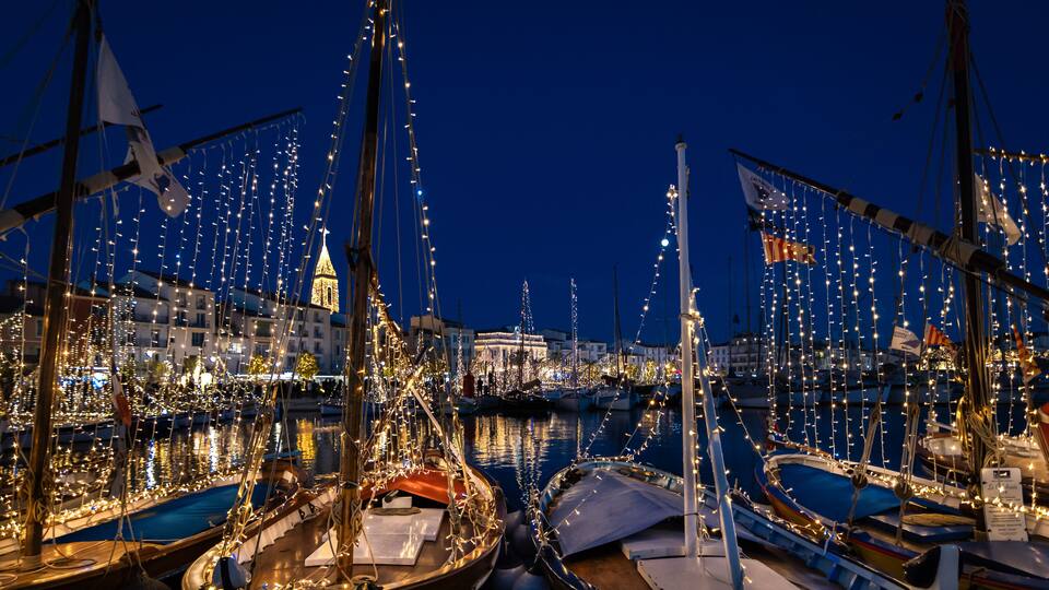 A marina at night with boats decorated with string lights and a prominent tower in the background.