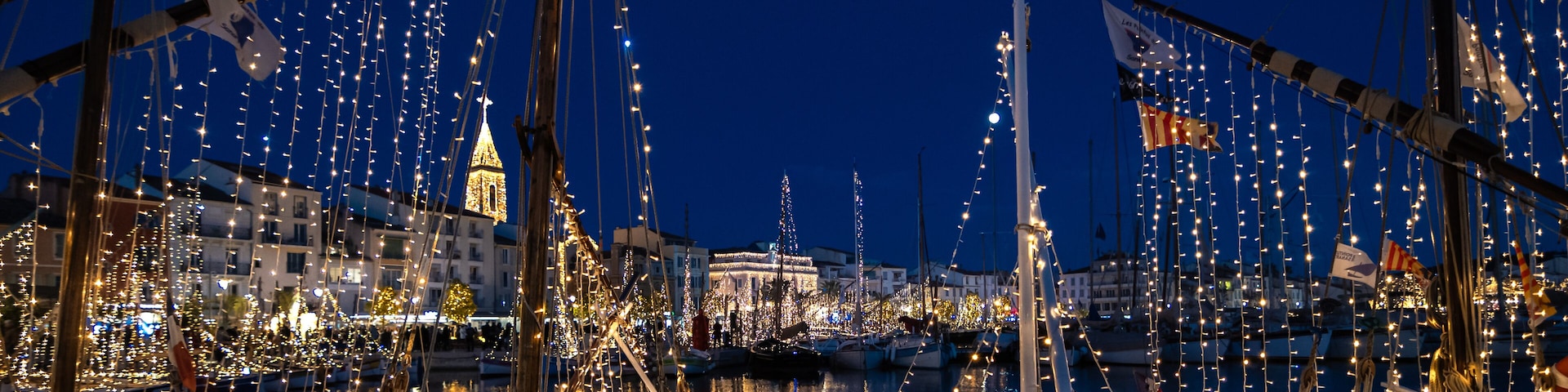 Un puerto deportivo de noche, con barcos decorados con luces de cadena y una torre destacada al fondo.