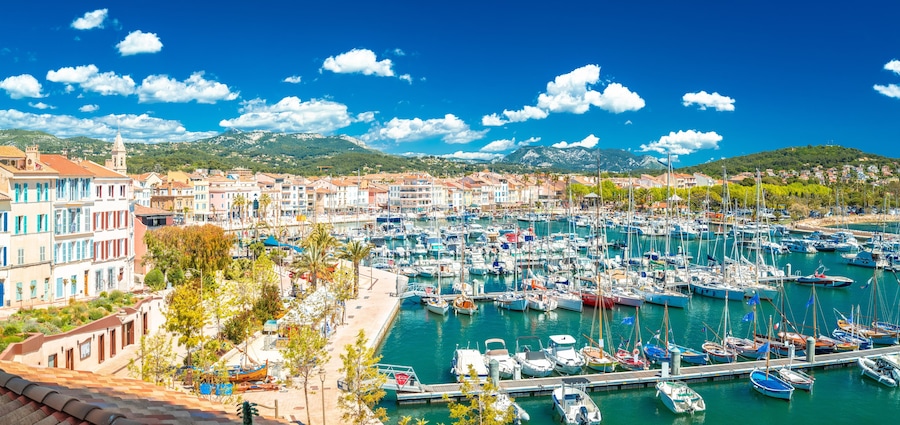 Un port de plaisance où sont amarrés de nombreux bateaux, une ville côtière aux bâtiments aux couleurs vives et, au loin, une chaîne de montagnes.