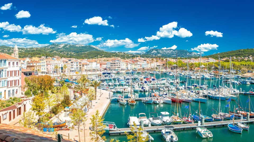A marina with numerous boats docked, a coastal town with colorful buildings, and a mountain range in the background.