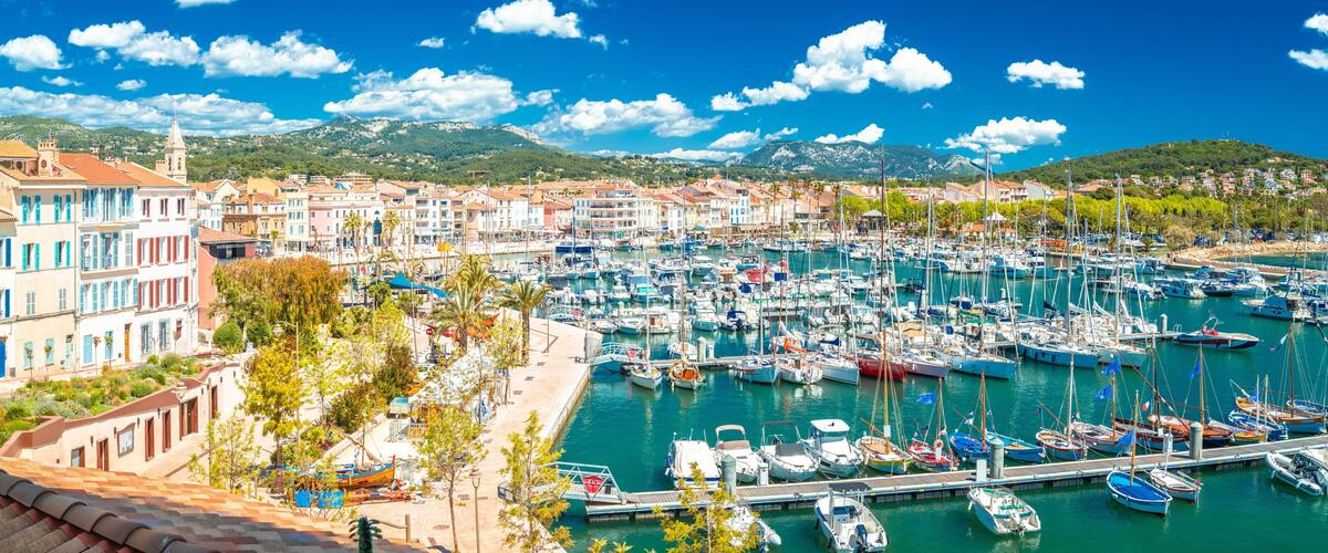 A marina with numerous boats docked, a coastal town with colorful buildings, and a mountain range in the background.