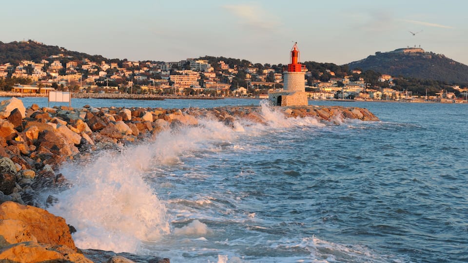 A coastal town with a prominent lighthouse, rocky shoreline, and a hill with buildings in the background.
