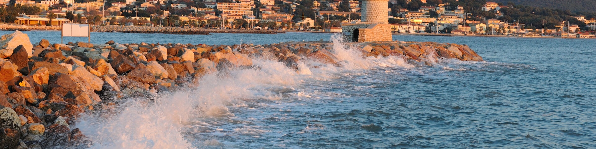 Un pueblo costero con un faro destacado, costa rocosa y una colina con edificios al fondo.