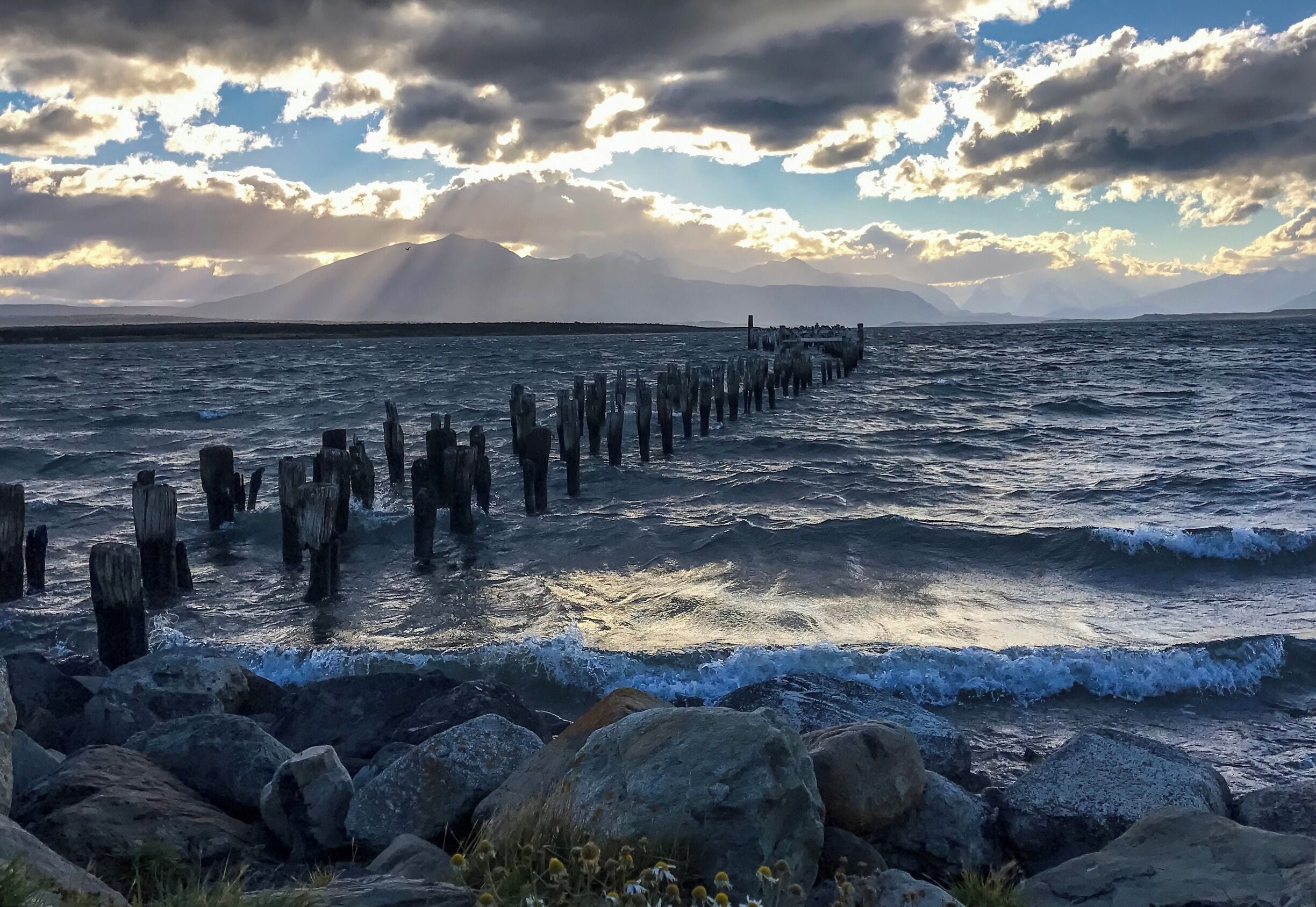 A rocky shoreline with a wooden pier extending into the sea, under a partly cloudy sky.